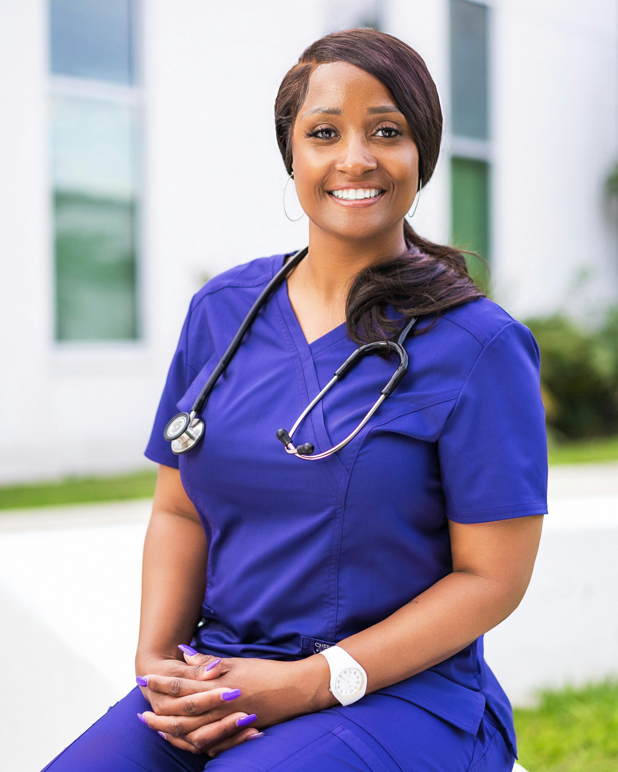 Portrait of a smiling female nurse in purple scrubs with a stethoscope, sitting outdoors.